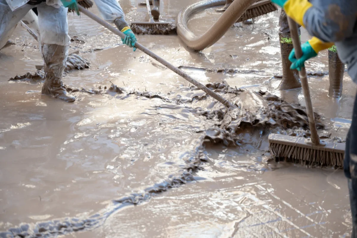 Workers wearing boots and gloves clearing muddy flood residue with brushes